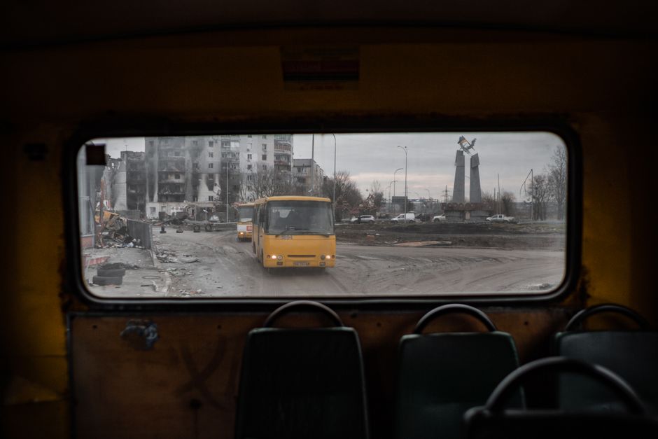 Imagem de uma rua urbana vista de dentro de um ônibus, mostrando uma paisagem de destruição e edifícios danificados, com uma estátua ao fundo.