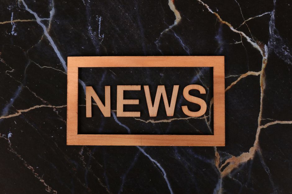 Imagem de um quadro de madeira com a palavra 'NEWS' em destaque, sobre um fundo de mármore preto com veios claros, simbolizando comunicação e atualização de notícias.