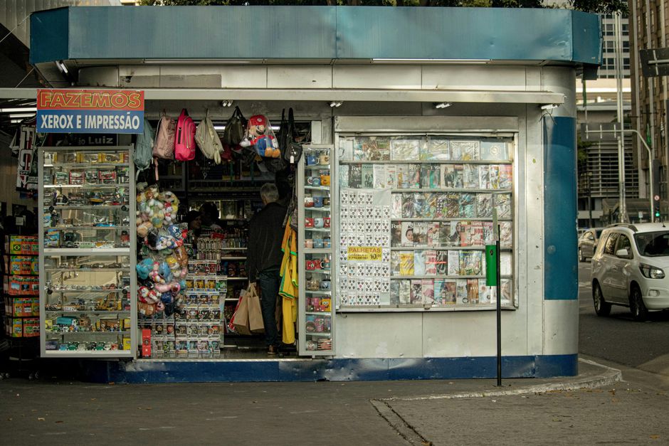 Imagem de uma banca de jornal e papelaria na rua, vendendo revistas, brinquedos e artigos diversos ao redor de uma cidade movimentada.