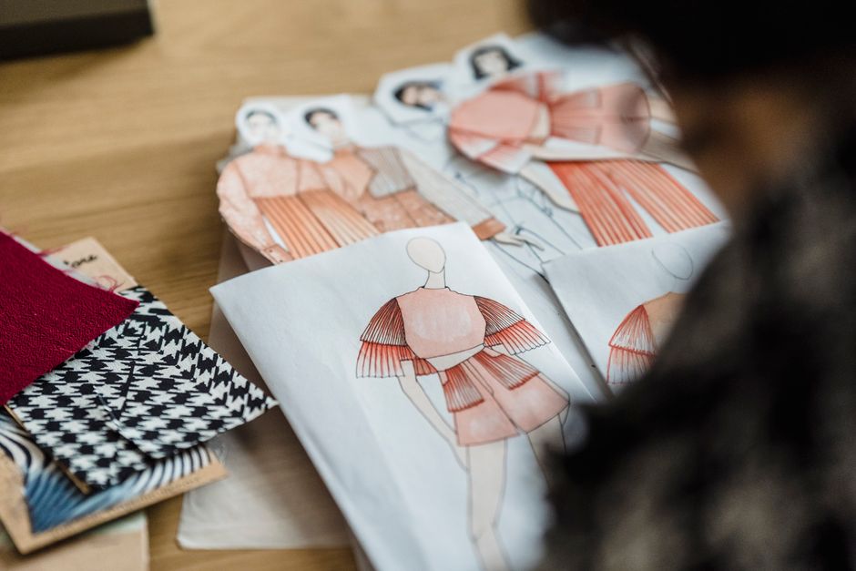 Pessoa analisando desenhos de roupas de moda, em um ambiente de estúdio ou ateliê de costura, com foco no processo criativo e de produção de vestuário.