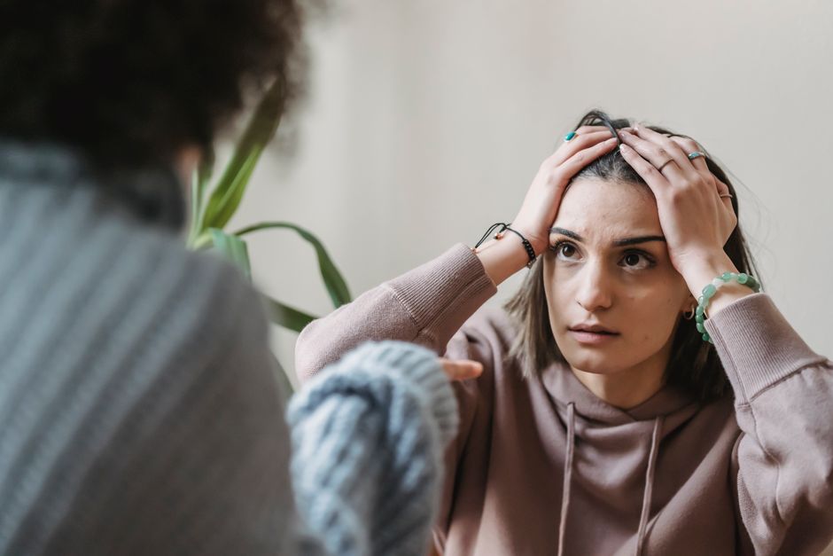 Jovem mulher com expressão de preocupação ou ansiedade, segurando a cabeça durante uma conversa, ilustrando uma situação de estresse emocional ou psicológico.