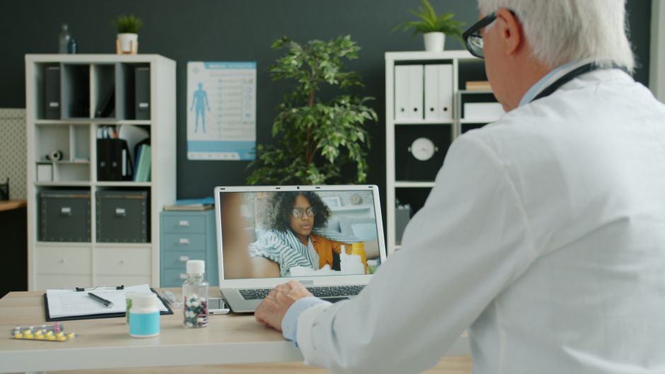 Profissional de saúde realizando uma consulta médica virtual com uma paciente usando computador em consultório moderno.