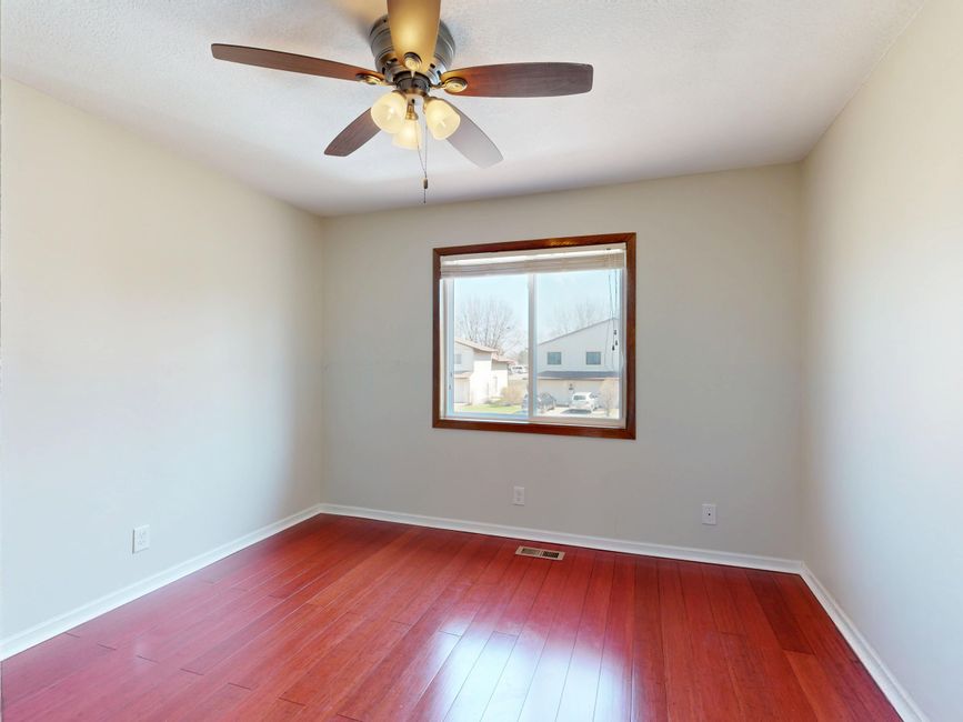 Quarto vazio com piso de madeira clara, parede branca, janela e ventilador de teto, pronto para decoração ou mudança.