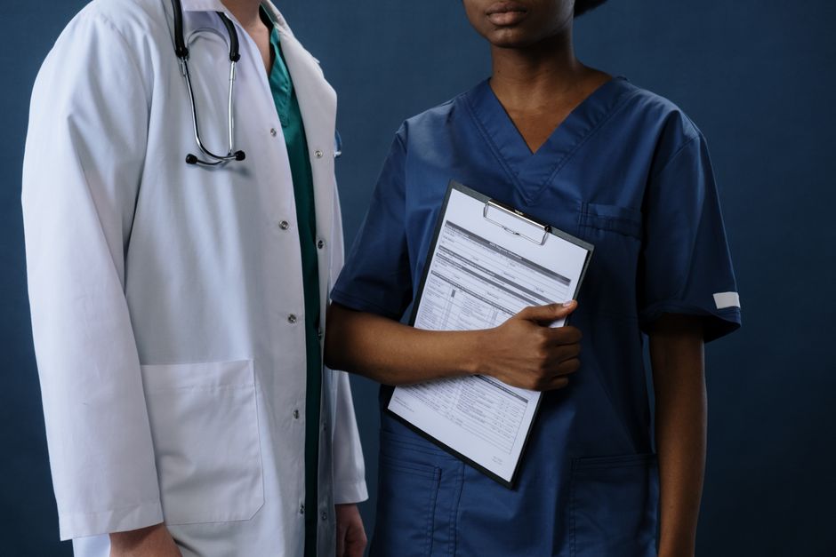 Jovem profissional da área de saúde segurando prontuário ao lado de médica, simbolizando trabalho em equipe e cuidados médicos.