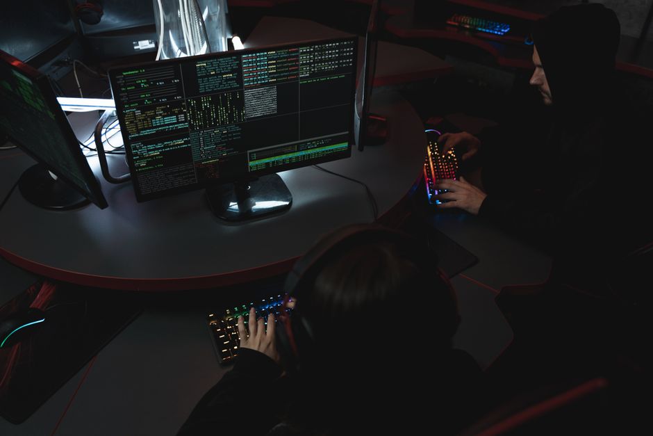 Imagem de hackers ou profissionais de segurança cibernética trabalhando em um ambiente de tecnologia avançada com múltiplos monitores e equipamentos de alta performance.
