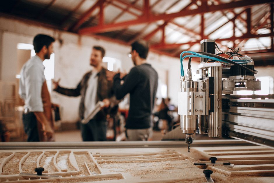 Imagem de um ambiente de produção industrial com máquinas de alta tecnologia e três pessoas discutindo ao fundo, destacando inovação e colaboração no setor manufatureiro.