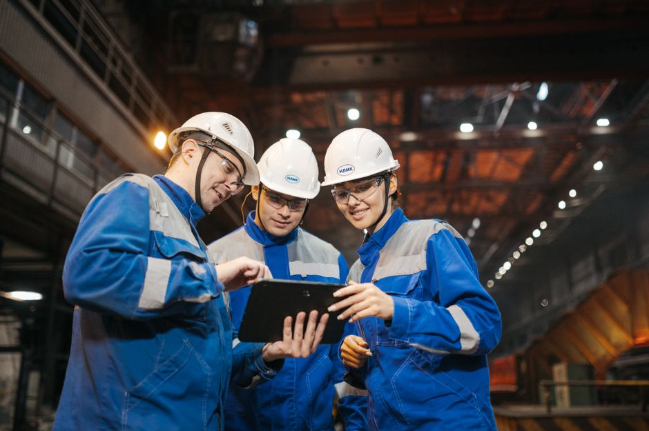 Três profissionais em fardamento de segurança industrial, discutindo informações em um tablet dentro de uma fábrica ou ambiente de produção.