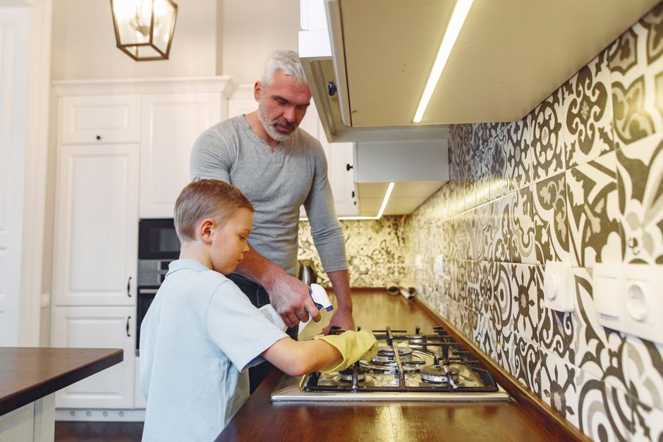 Um homem ajudando uma criança a limpar ou consertar o fogão na cozinha de casa, reforçando valores de convivência e aprendizado familiar.