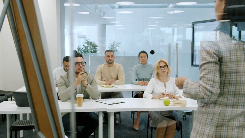 Reunião de equipe em um ambiente corporativo, com uma pessoa apresentando informações aos colegas atentos ao tema de gestão ou treinamento empresarial.