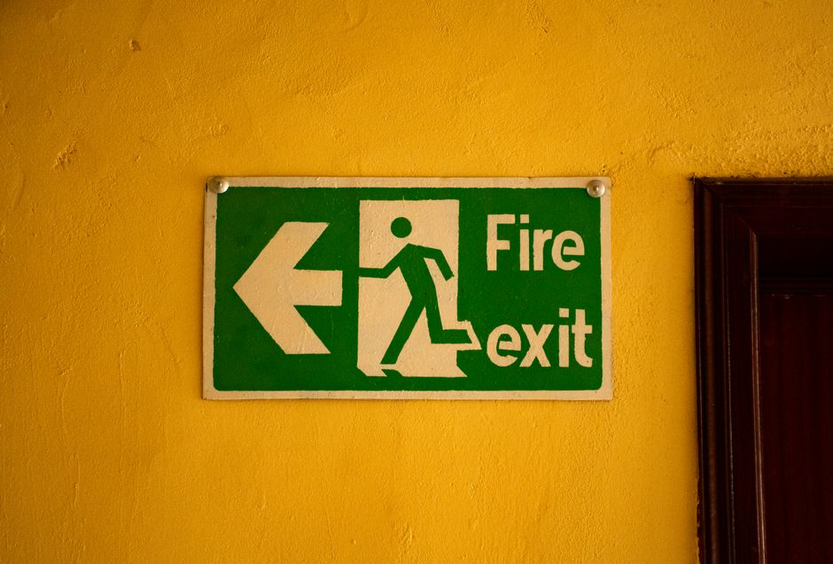 Placa de sinalização indicando a saída de emergência contra incêndio em um ambiente interno com parede amarela ao fundo.