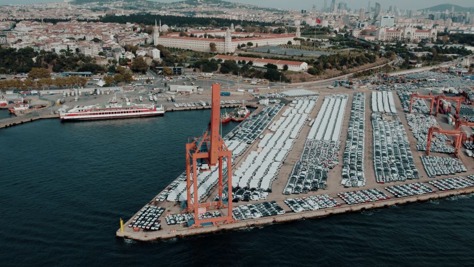 Vista aérea de um porto de carga com inúmeros veículos estacionados, mostrando a movimentação na região portuária e industrial da cidade.