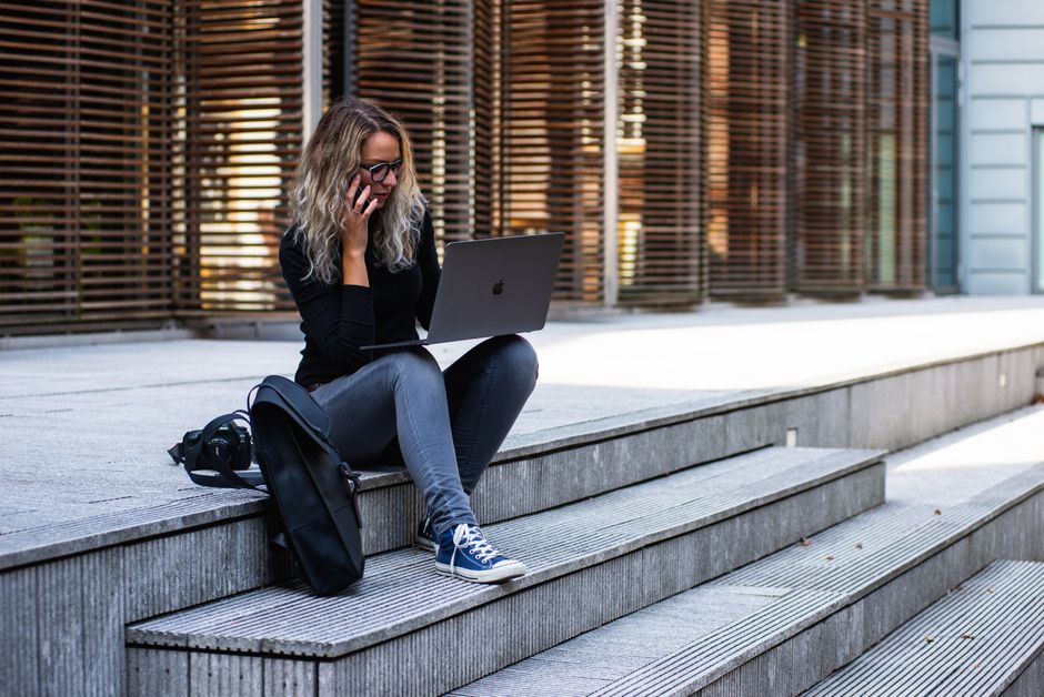 Mulher usando laptop e celular enquanto está sentada em degraus na área externa, representando trabalho remoto e conectividade.