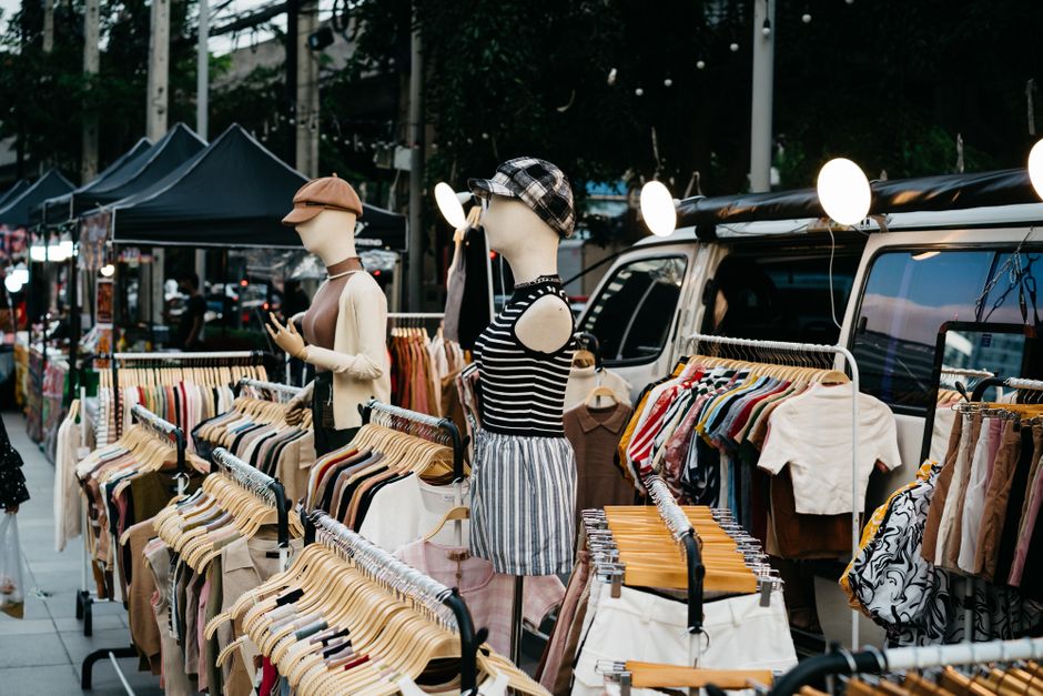 Imagem de uma feira ao ar livre com manequins vestidos com roupas de verão, destacando o comércio de moda e acessórios em espaço externo.