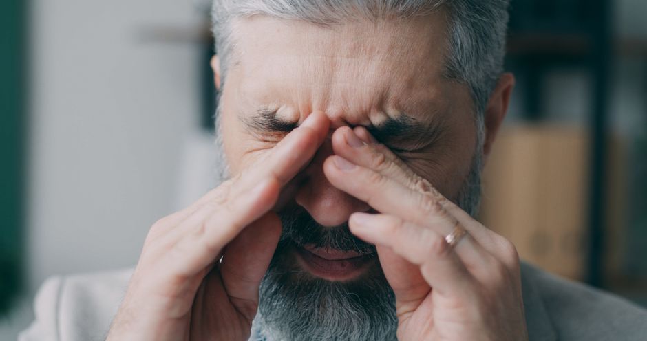 Homem com expressão de estresse ou dor, segurando o rosto com as mãos, em um ambiente interno.