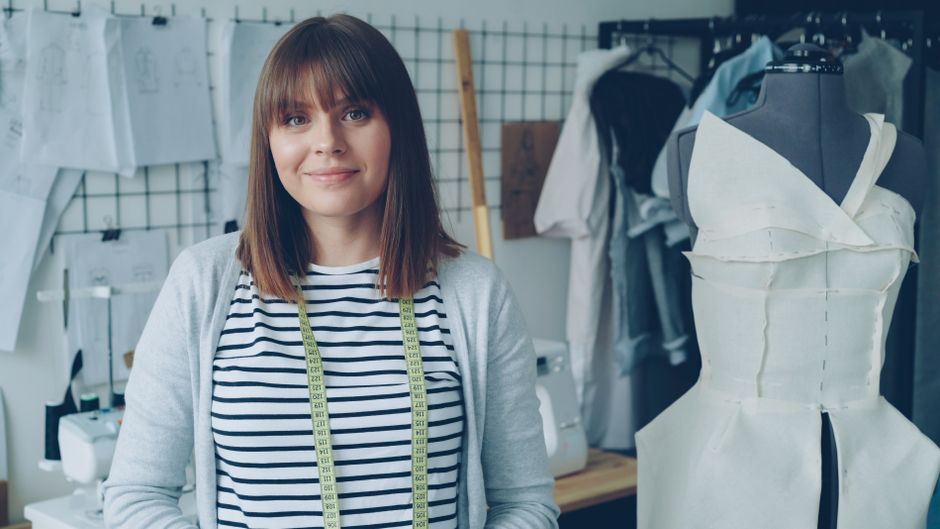Jovem estilista ou costureira em seu ateliê, cercada por manequins e moldes de roupas, em ambiente criativo de moda e confecção.