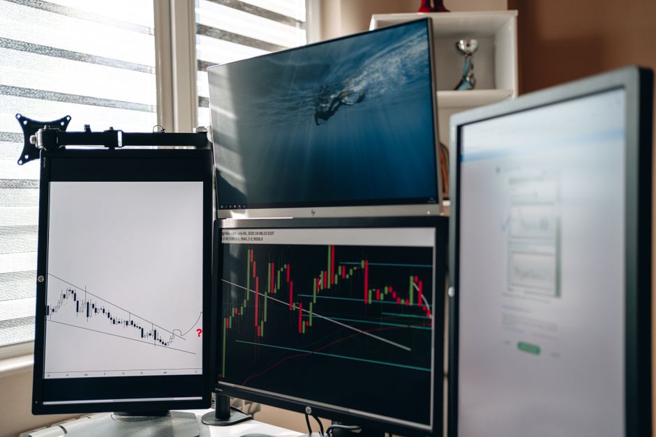 Imagem de estação de trabalho com múltiplos monitores exibindo gráficos financeiros, ideal para um artigo sobre análise de investimentos ou trader profissional.