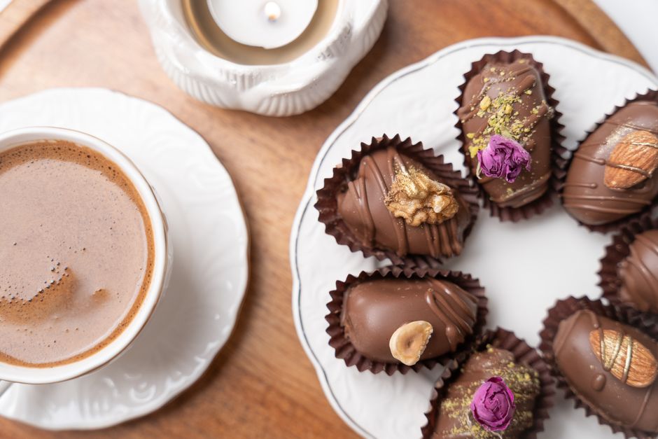 Imagem de uma xícara de chocolate quente ao lado de uma variedade de chocolates decorados, sugerindo um momento de pausa ou lazer com doces.