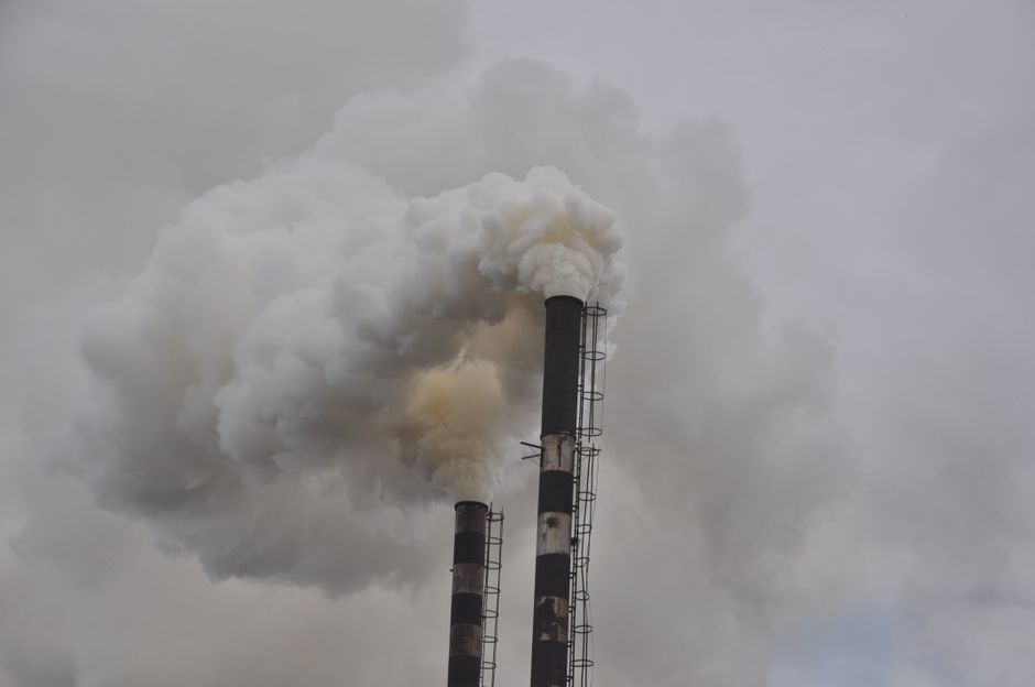Imagem de duas torres de fumaça emitindo poluição industrial contra um céu cinzento, simbolizando questões ambientais e sustentabilidade.