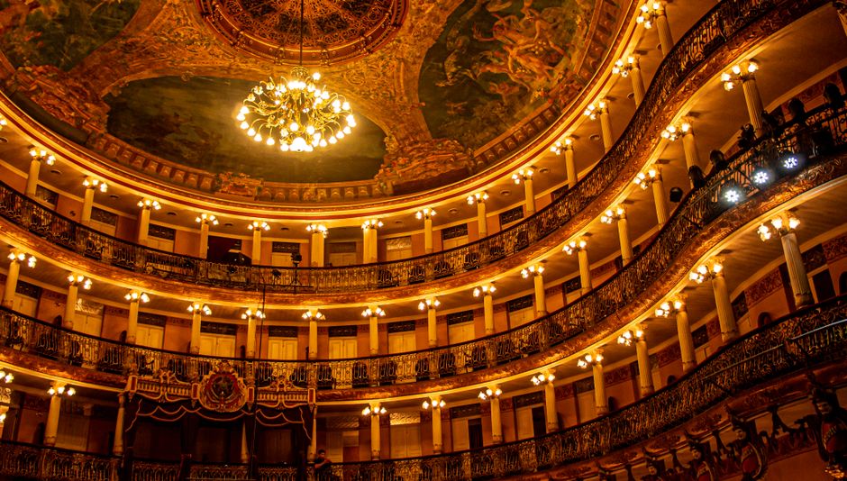 Interior de um teatro ou teatro antigo com iluminação de candelabros dourados e teto decorado com pinturas artísticas.