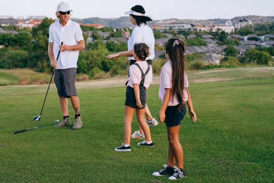 Adulto ensinando crianças a jogar golfe em um campo aberto, promovendo educação esportiva e lazer ao ar livre.