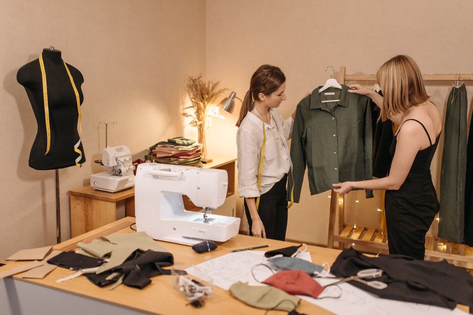 Duas mulheres avaliam uma camisa em um brechó ou loja de roupas, enquanto trabalham na confecção ou venda de roupas sustentáveis.