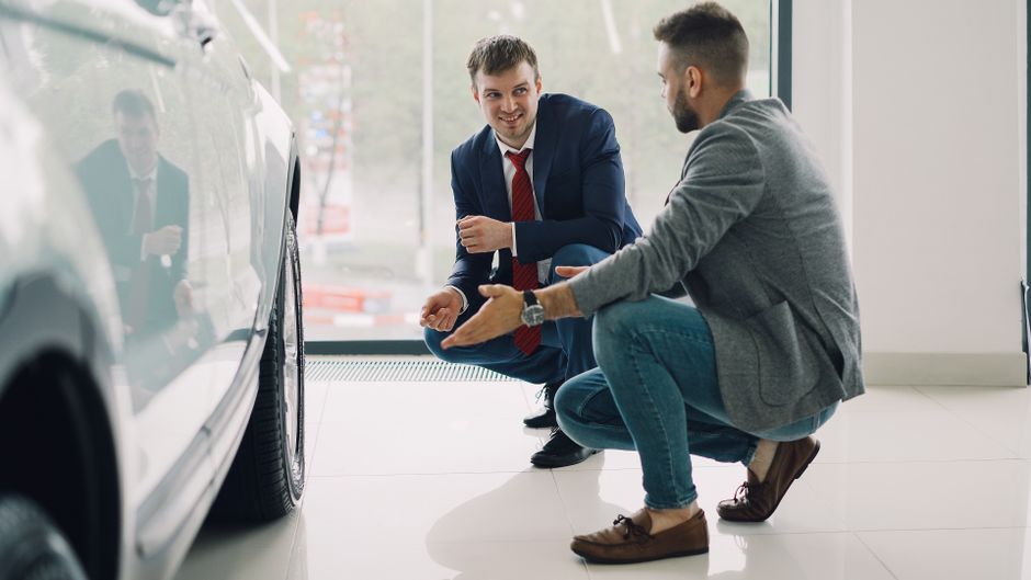 Dois vendedores conversando com cliente sobre detalhes de um carro novo em uma concessionária moderna.