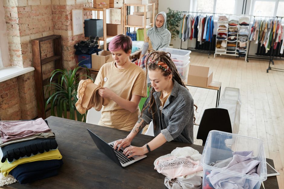 Mulheres organizando roupas e acessórios em uma loja de roupas ou espaço de vendas, focando no processo de gestão de estoque ou visual merchandising.
