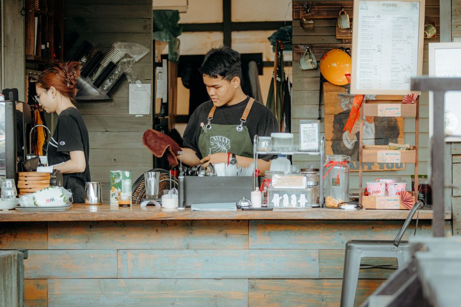 Duas pessoas atendendo clientes em uma cafeteria com ambiente rústico de madeira, focando no serviço de alimentação e bebidas artesanais.