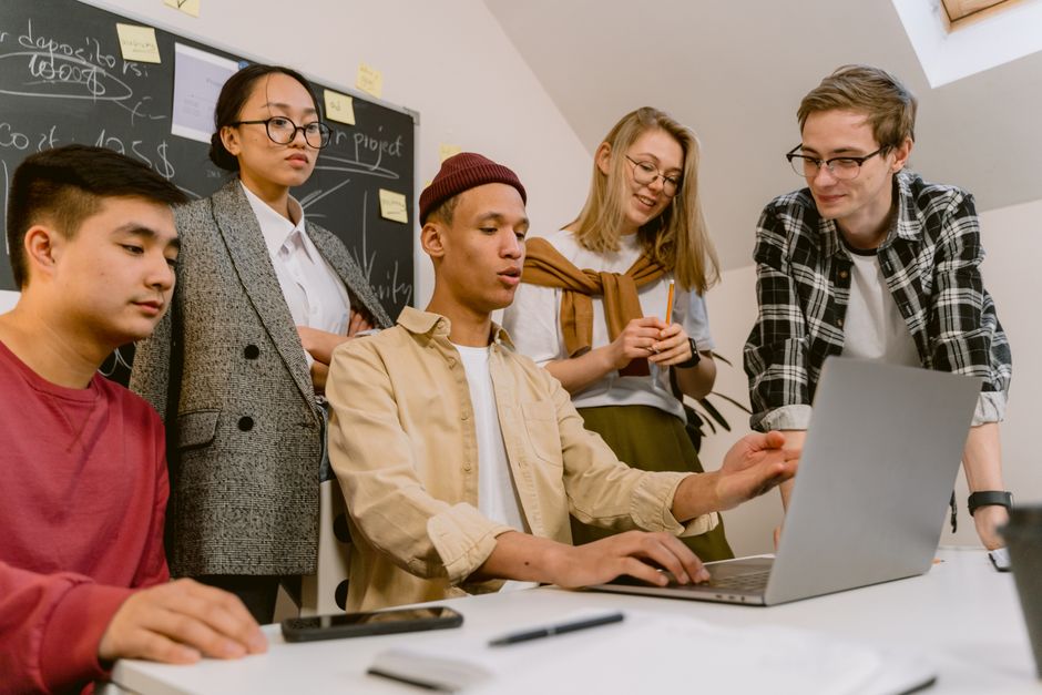 Grupo de jovens profissionais colaborando em uma reunião de equipe em um ambiente de escritório moderno com quadro branco ao fundo.
