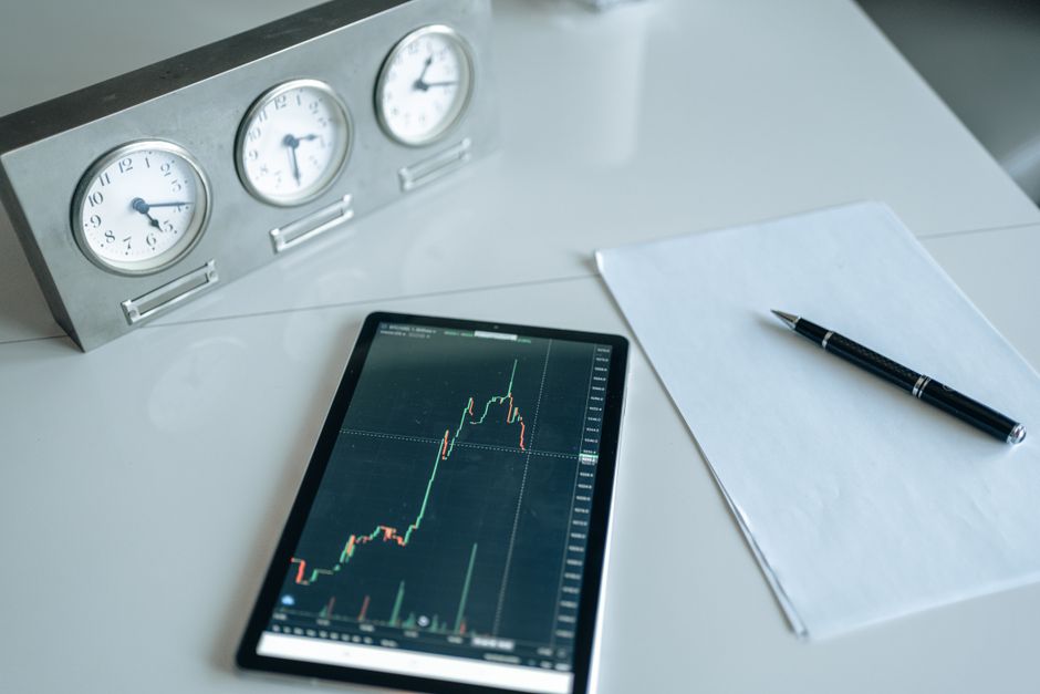 Imagem de um ambiente de trabalho com foco em análise financeira, destacando um gráfico de investimentos em um tablet, acompanhado de uma caneta, uma folha de papel e um relógio de três mostradores na mesa.