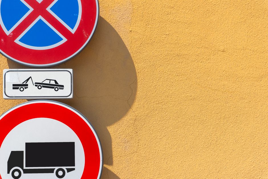 Placas de trânsito indicando restrições de estacionamento e transporte em frente a uma parede amarela.