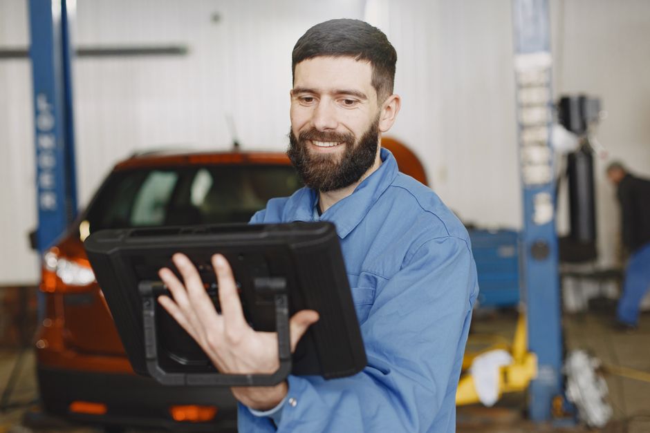 Homem sorridente trabalhando na oficina mecânica, usando tablet para verificar informações técnicas ou realizar inspeções.