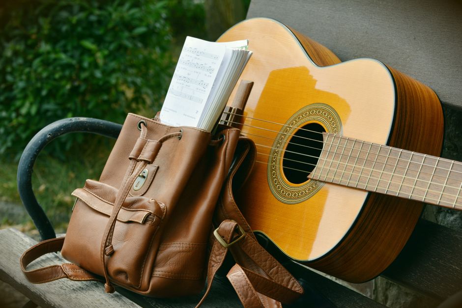 Imagem de uma guitarra acústica apoiada em um banco ao lado de uma bolsa de couro com um caderno de partituras, em ambiente externo com vegetação ao fundo.