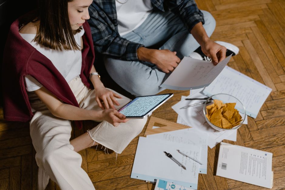 Pessoas estudando e revisando materiais acadêmicos em um ambiente descontraído com documentos e lanche ao redor.