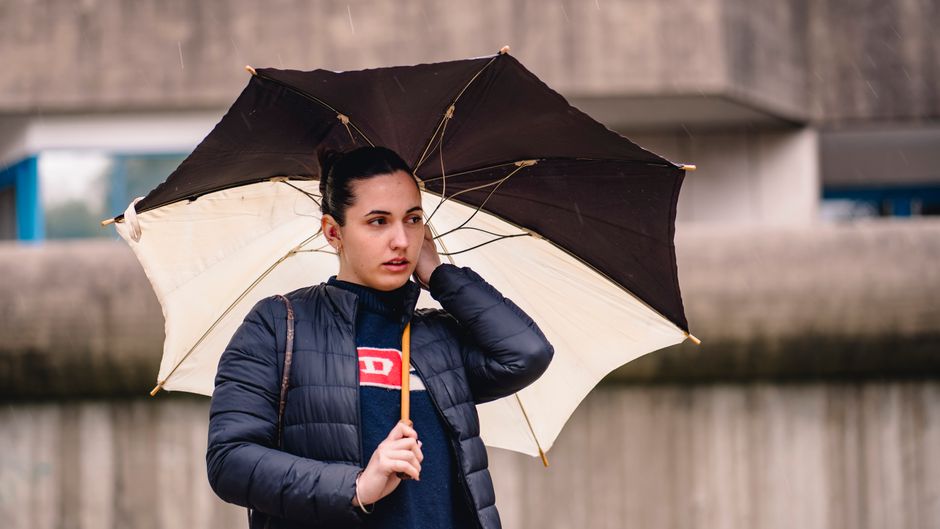 Mulher de cabelo escuro usando jaqueta e segurando guarda-chuva, com expressão pensativa ao ar livre sob chuva.