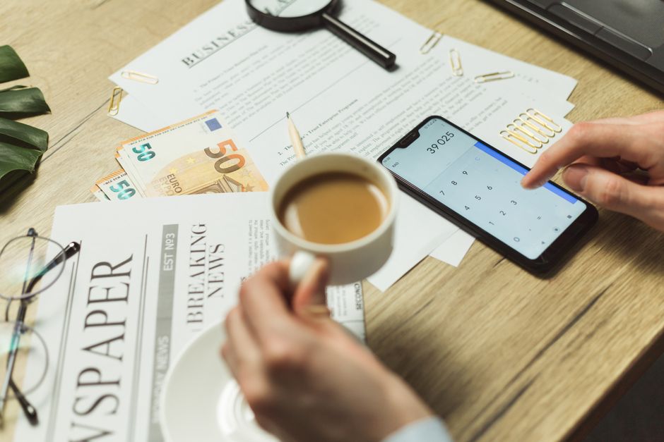 Imagem de uma mesa de trabalho com documentos, dinheiro em euros, celular com aplicativo de cálculo, e uma pessoa segurando uma xícara de café, representando temas de planejamento financeiro e negócios.