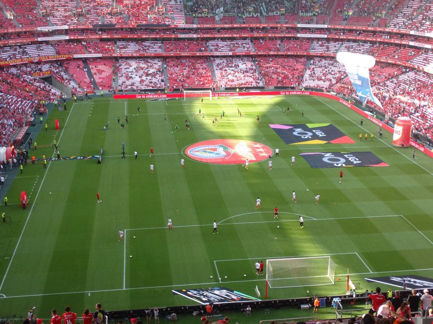 Visão geral de um estádio de futebol durante o aquecimento antes do jogo, com foco na atmosfera esportiva e torcida animada.
