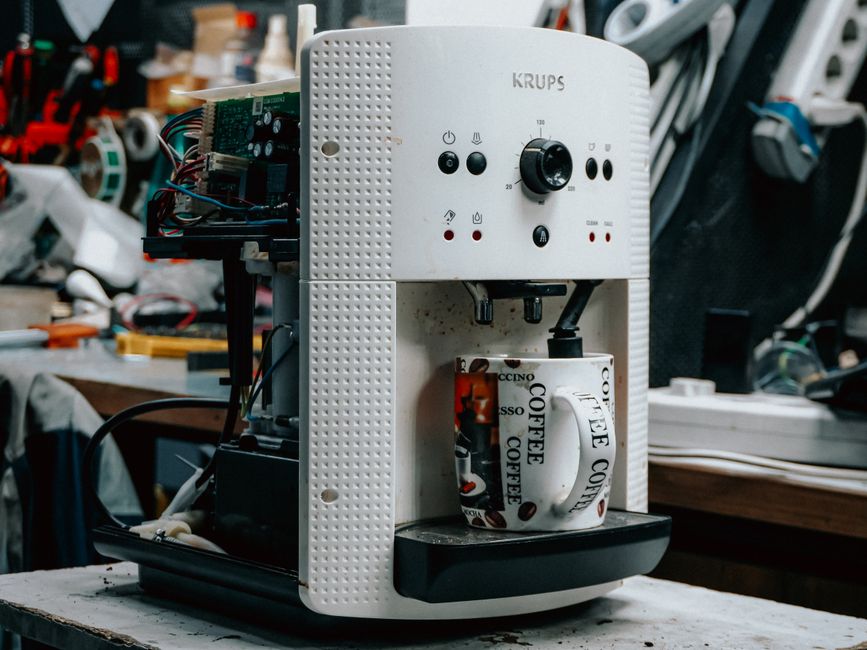 Imagem de uma máquina de café automática desmontada, destacando componentes eletrônicos e mecânicos, em ambiente de oficina ou laboratório de tecnologia.