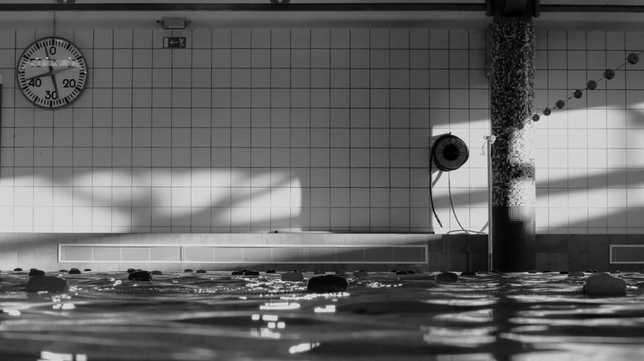 Imagem de uma piscina coberta, vista do nível da água, com destaque para o relógio na parede e elementos arquitetônicos do espaço.