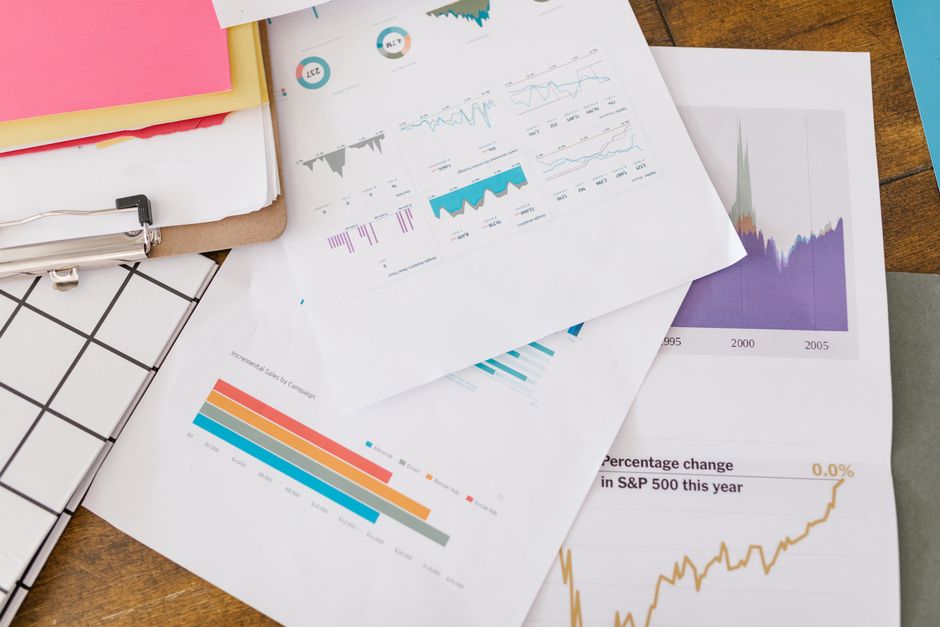 Grupos de relatórios e gráficos de desempenho financeiro sobrepostos em uma mesa de trabalho, representando análise de dados e planejamento financeiro.