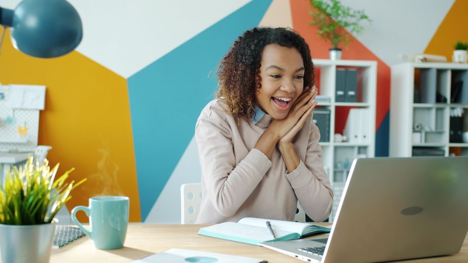 Pessoa celebrando uma conquista enquanto trabalha em um ambiente de escritório colorido, com laptop e materiais de estudo à frente.