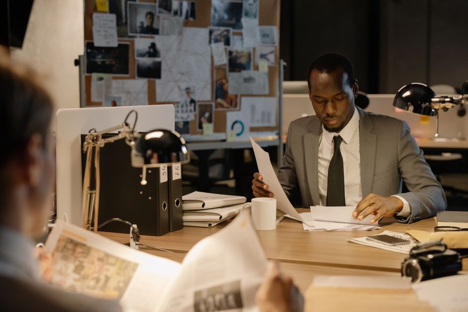 Homem de terno analisando documentos em um ambiente de trabalho com quadro de avisos ao fundo, sugerindo uma atividade de investigação ou planejamento estratégico.