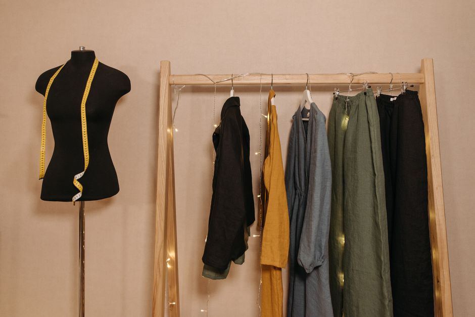 Imagem de um manequim com fita métrica em volta do pescoço ao lado de um cabideiro com roupas, simbolizando moda, confecção ou estilismo.