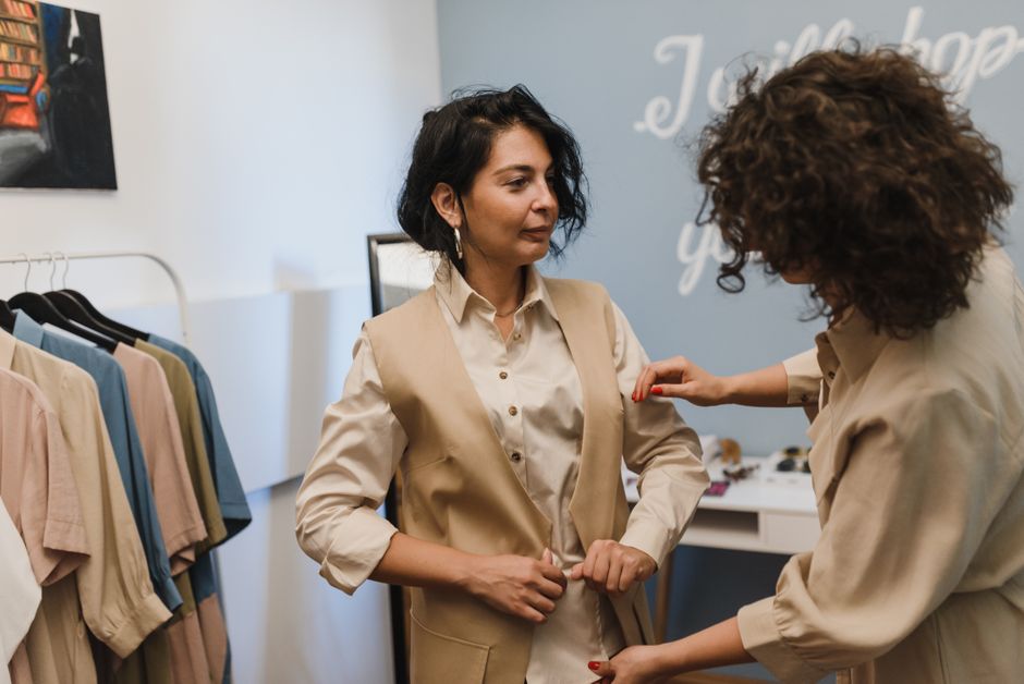 Duas mulheres em uma conversa amistosa dentro de uma loja de roupas, enquanto uma delas ajusta a blusa da outra, destacando o ambiente de comércio e relacionamento com clientes.