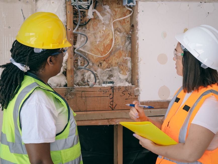 Duas engenheiras discutindo ou avaliando uma estrutura em uma obra em andamento, com uso de equipamentos de segurança e anotações técnicas.