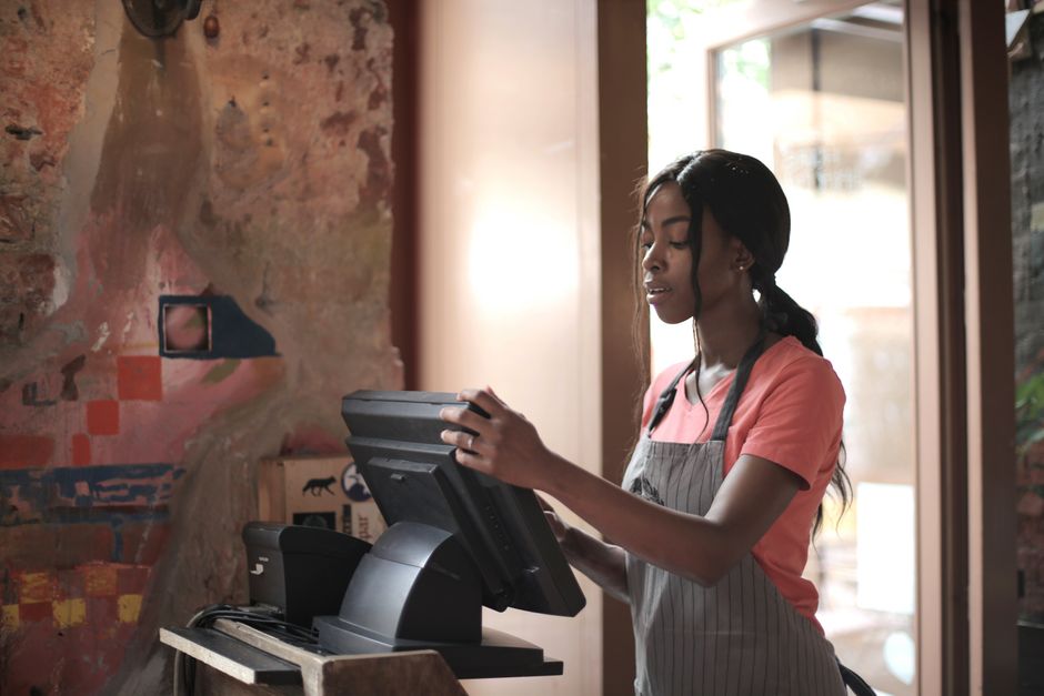 Jovem atendente operando o sistema de checkout em uma cafeteria com ambiente acolhedor e decorado com parede de tijolos expostos.