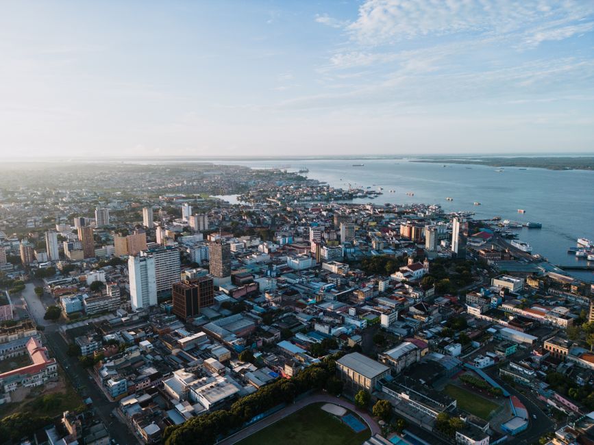 Vista aérea de uma cidade litorânea sob um céu ensolarado, destacando a combinação de ambientes urbanos e costeiros.