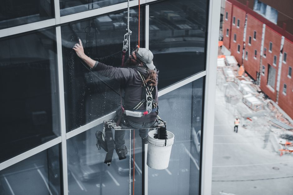Profissional de limpeza fazendo manutenção em janelas de um edifício comercial alto, destacando cuidados e técnicas de limpeza em altura.