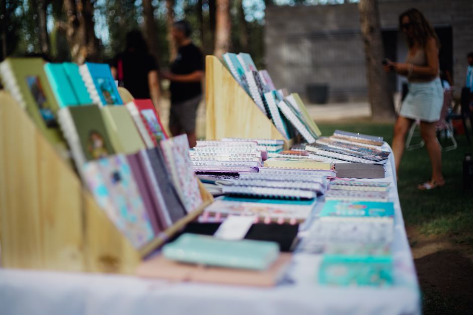 Imagem de uma banca ao ar livre exibindo livros e cadernos coloridos, com pessoas caminhando ao fundo em um ambiente de natureza.