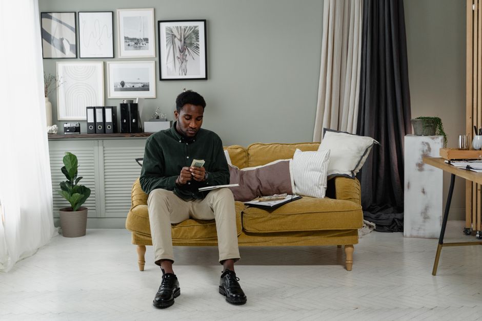 Homem sentado em um sofá amarelo, olhando para o telefone em um ambiente de casa com decoração moderna e acolhedora.
